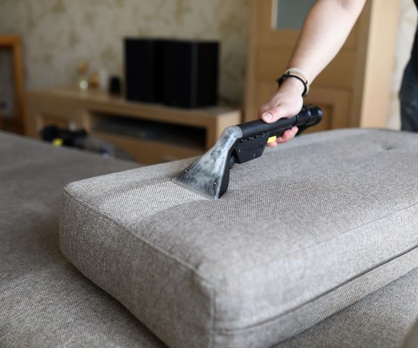 Un homme de ménage nettoie un canapé à la main avec un aspirateur.